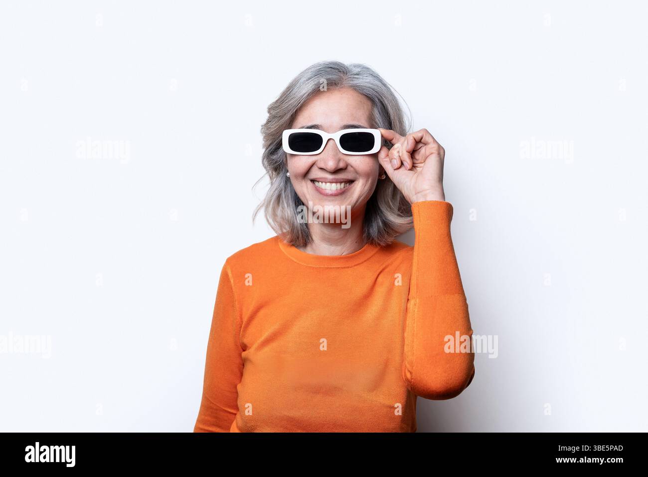 Eine asiatische chinesische Seniorin mit grauem Haar, die gerne eine stilvolle weiße Sonnenbrille und einen orangefarbenen Pullover trägt und ein Gefühl von Selbstvertrauen und Freude ausstrahlt Stockfoto