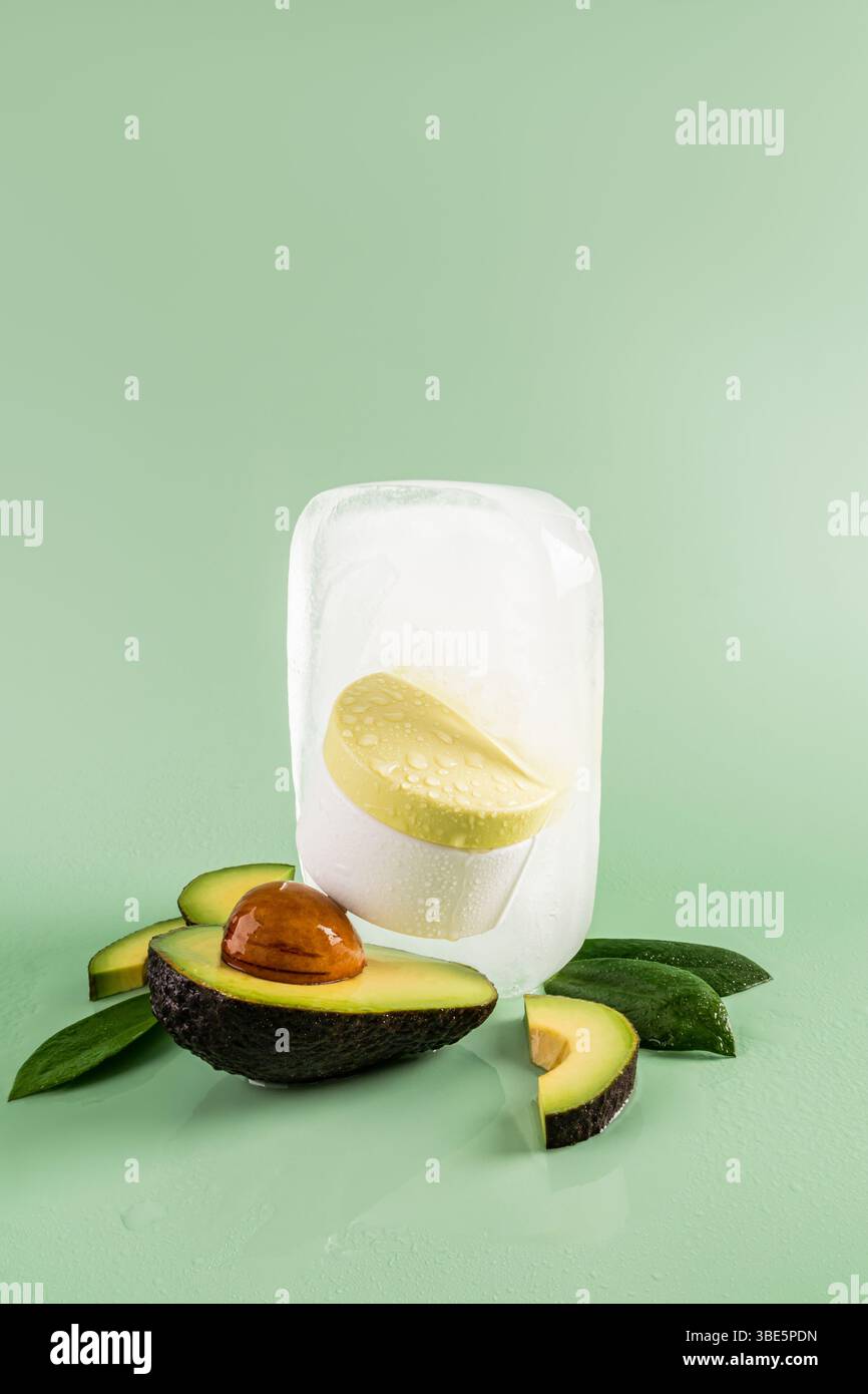 Frische Avocadoblätter und Hautpflegeprodukte in einem klaren Eisblock, umgeben von geschnittenen Avocados auf einem glatten grünen Hintergrund, die eine lebendige, raffinierte Atmosphäre schaffen Stockfoto
