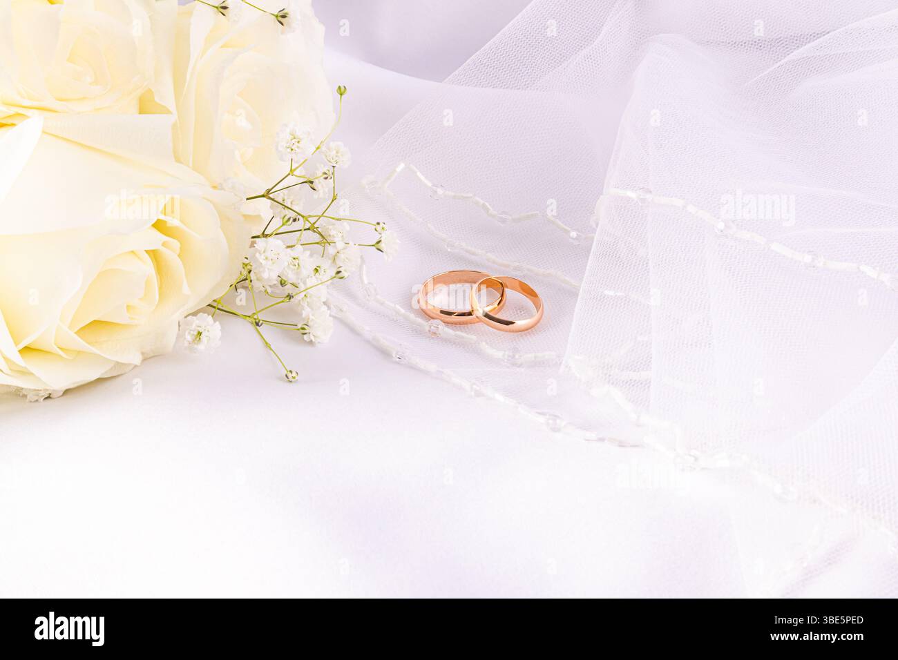 Zwei elegante Eheringe liegen sanft zwischen zarten Creme-Rosen und dem Atem des Babys auf Stoff und symbolisieren Einheit und Liebe. Die weichen Texturen erinnern an roma Stockfoto