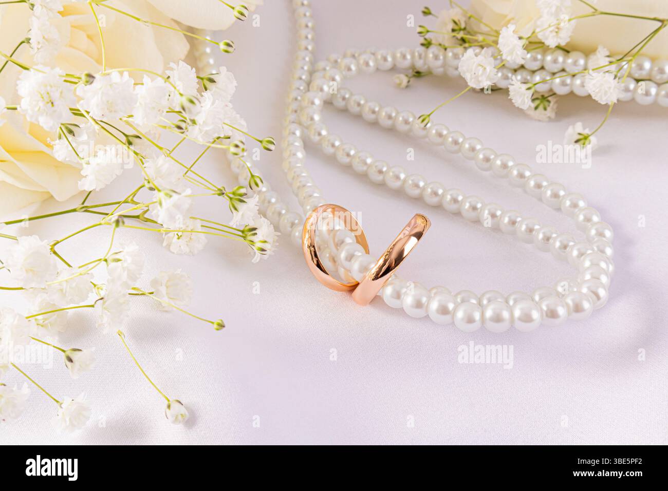 Elegante Eheringe gepaart mit zarten Blumen und einer Perlenschnur auf einem weichen weißen Hintergrund, die Liebe und Einheit in einer schönen, ruhigen Atmosphäre symbolisieren Stockfoto