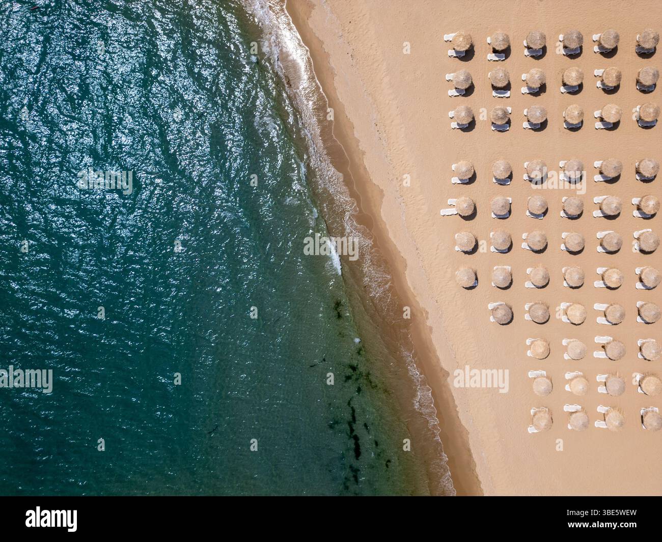 Von oben aus blickt man auf einen Sandstrand mit Strohschirmen und Liegen, die in Reihen angeordnet sind, und Wellen, die sanft an Land waschen. Sommerferienkonzept Stockfoto