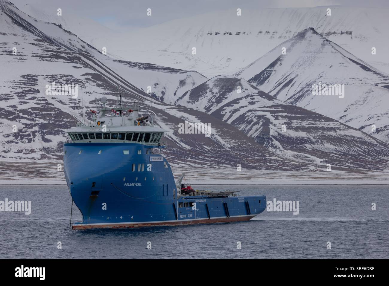 Hellblaues Svalbard Regierungsschiff Polarsyssel vor Anker in der Nähe der verschneiten Bergküste von Svalbard, Norwegen, im Frühling. Stockfoto