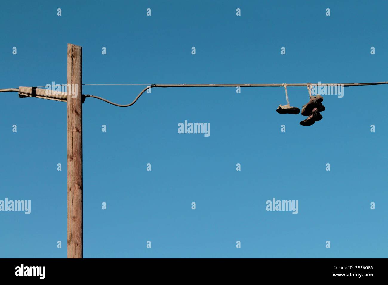 Vier gebrauchte Schuhe hängen an den Schnürsenkeln von einer Stromleitung, die an einem Holzpfosten befestigt ist, gegen einen klaren blauen Himmel. Da ist niemand. Stockfoto