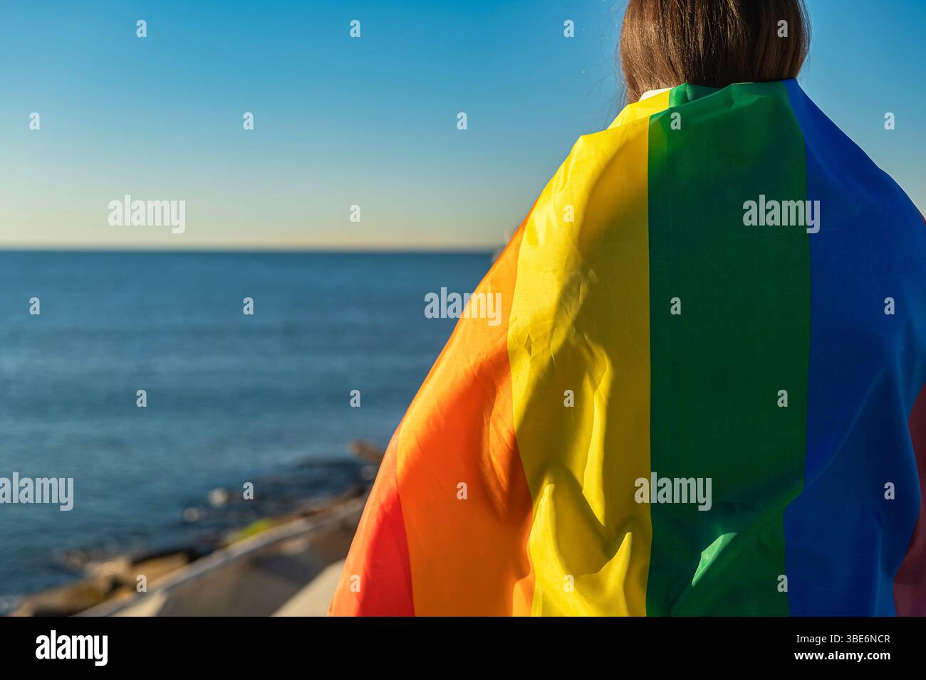 Horizontales Porträt einer jungen Frau, die in eine LGTBI-Flagge gehüllt ist, halblang und von einem Pier aus auf das Meer blickt. Das natürliche Licht leuchtet und leuchtet Stockfoto