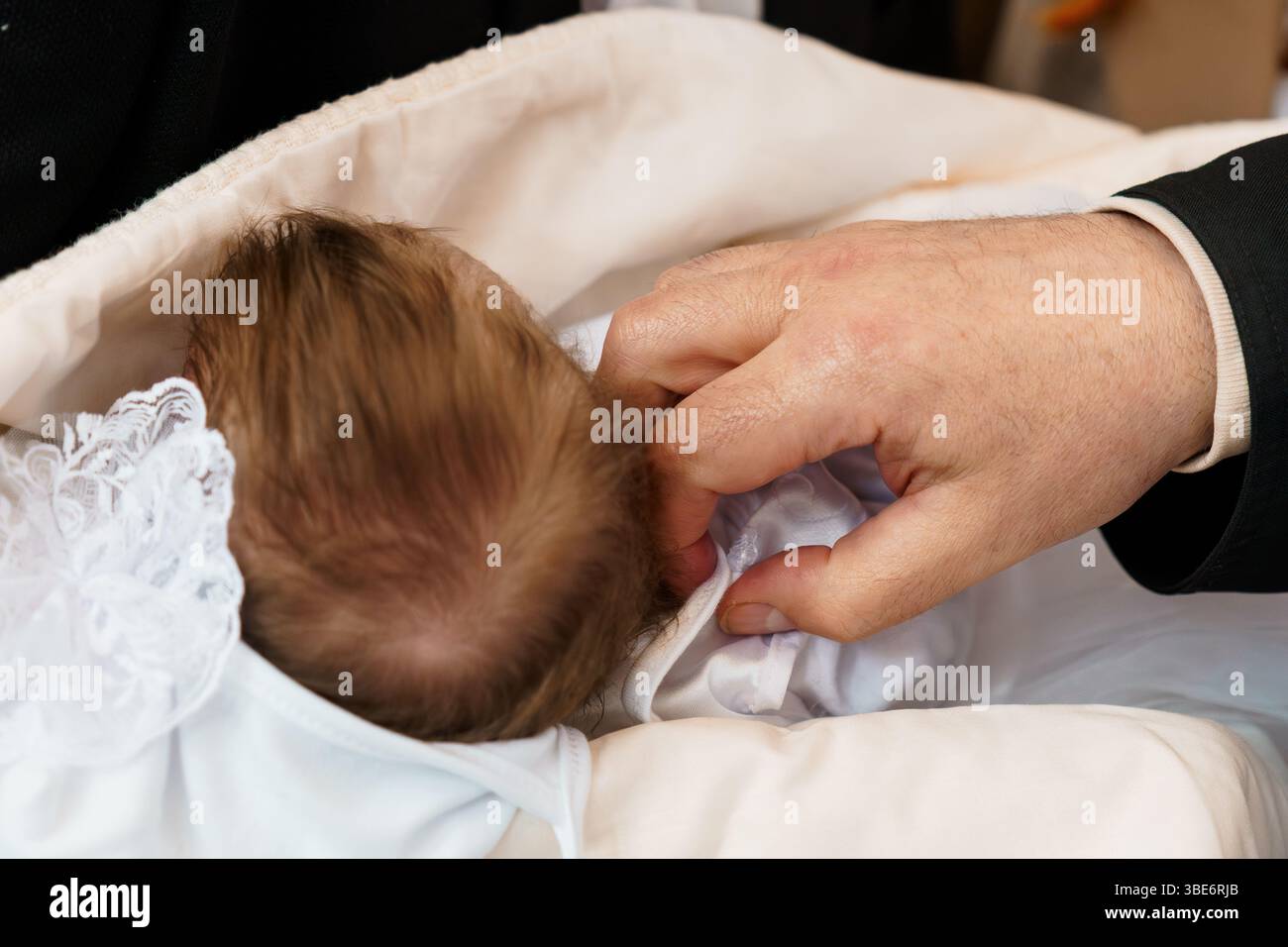 Ein zärtlicher Moment während der Taufzeremonie eines Babys, in dem die Hände eines Priesters den Kopf eines Säuglings sanft berühren, das in eine weiße Decke und Spitze gewickelt ist. Stockfoto
