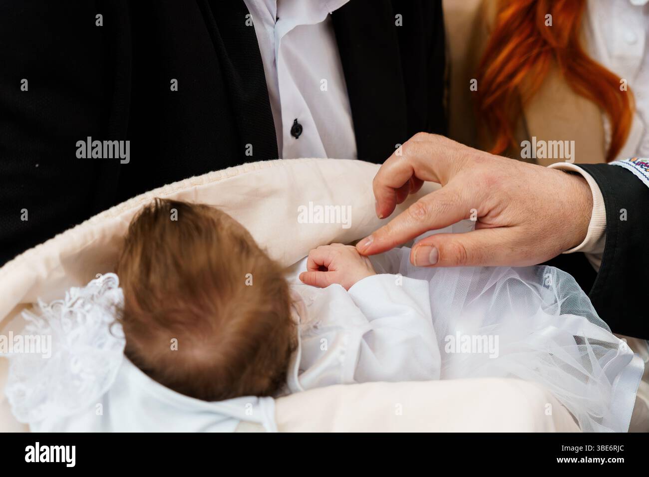 Ein zärtlicher Moment während der Taufzeremonie eines Babys, bei dem ein Mann sanft die Hand eines Säuglings berührt, das in einem weißen, mit Spitze besetzten Korb liegt, mit blu Stockfoto