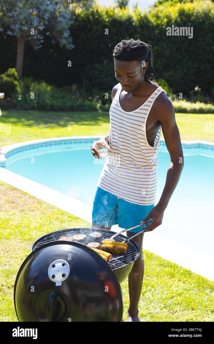 Grillender afroamerikanischer Mann in Badehosen, der Burger umwirft, Mais am Pool, mit Drink und Zange Stockfoto