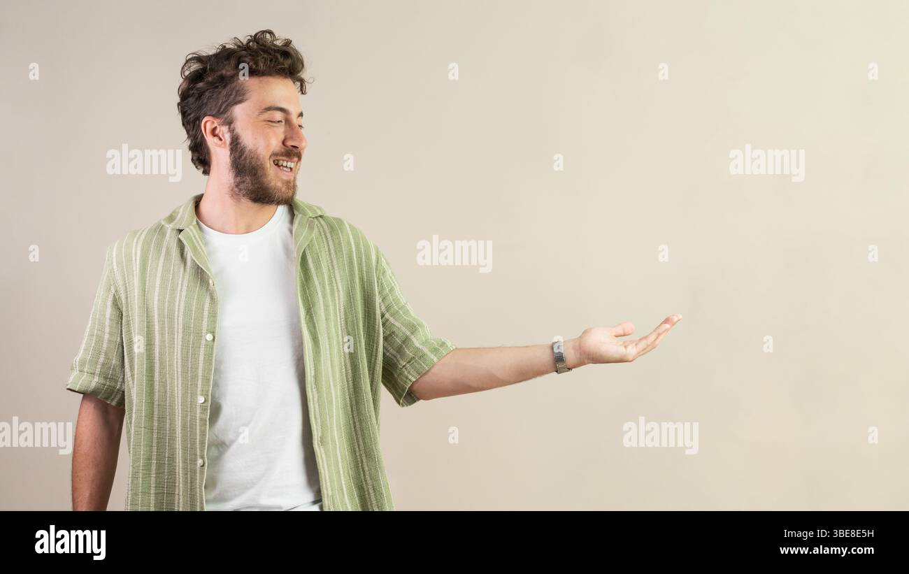Lächelnder junger Mann in grünem Hemd, der mit der Hand Gesten und einladendem Ausdruck zeigt, ideal für Produktplatzierung oder Werbedesign. Stockfoto
