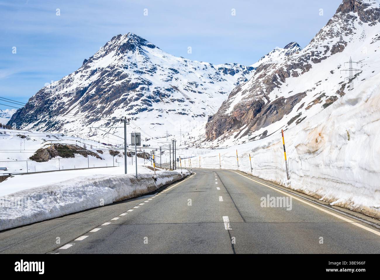 Leere kurvenreiche Straße, die an einem teilweise bewölkten Frühjahrstag entlang einer Bahn durch einen verschneiten Bergpass in den Schweizer Alpen führt Stockfoto