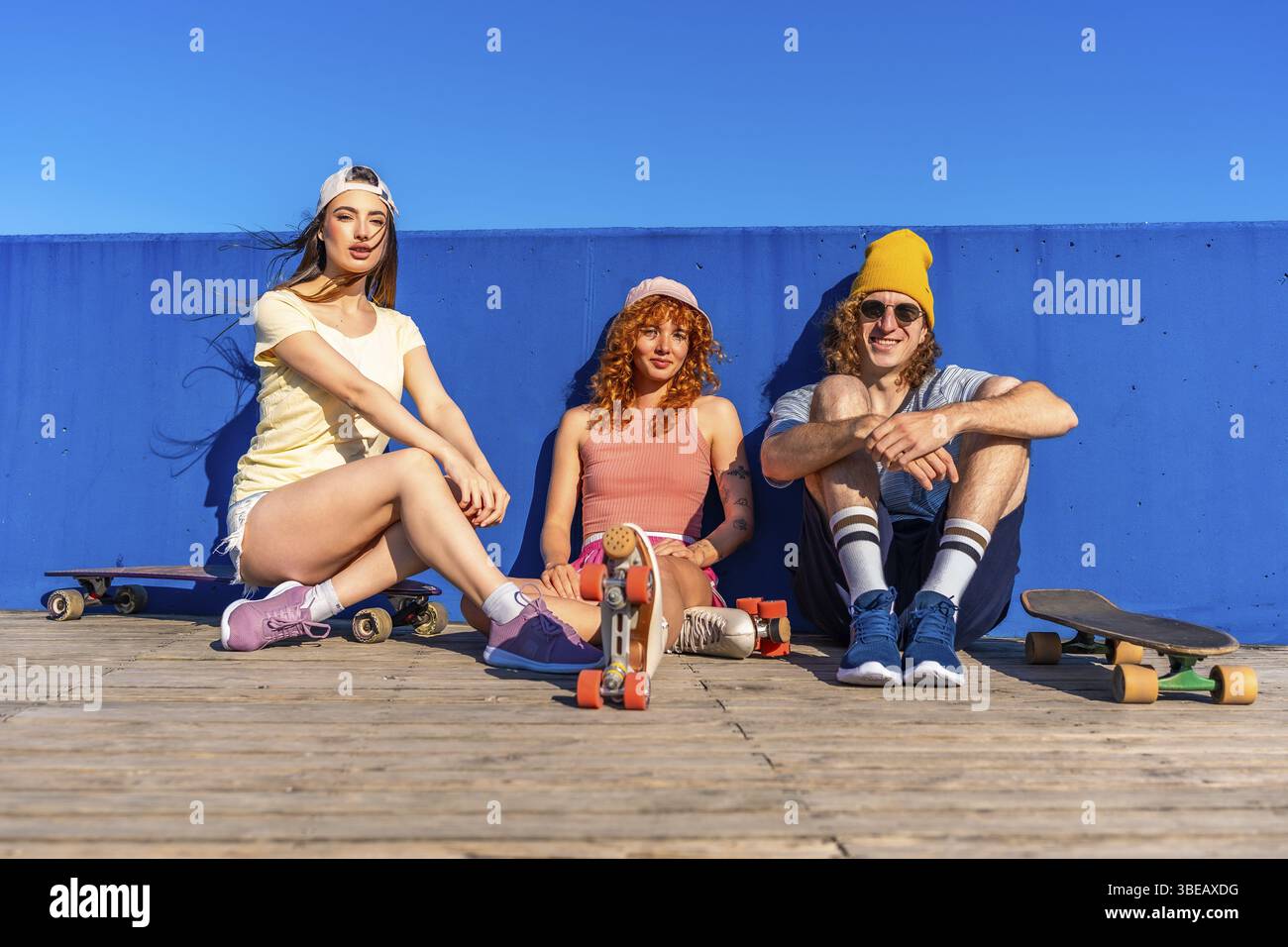 Drei junge Freunde entspannen sich an einem sonnigen Tag mit Skateboards und zeigen einen lebendigen und aktiven Lebensstil vor einem hellblauen Hintergrund Stockfoto