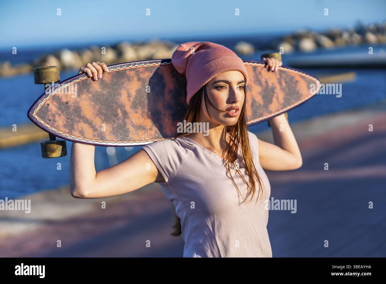 Eine junge Frau, die den Sonnenuntergang am Meer genießt, ein Longboard auf den Schultern hält und Freiheit und einen trendigen, aktiven Lebensstil verkörpert Stockfoto