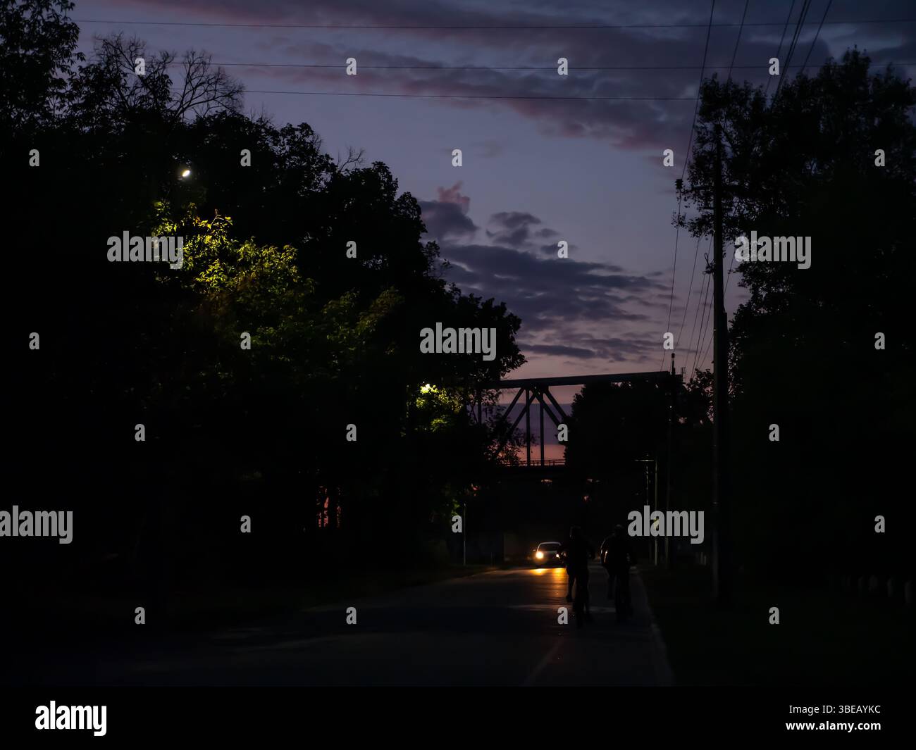 Radfahrer fahren mit dem Fahrrad auf einem Fahrradweg in Richtung einer Brücke und einem wunderschönen farbenfrohen Sonnenuntergang. Auto mit eingeschaltetem Scheinwerfer, Bäume mit Lampen hervorgehoben Stockfoto
