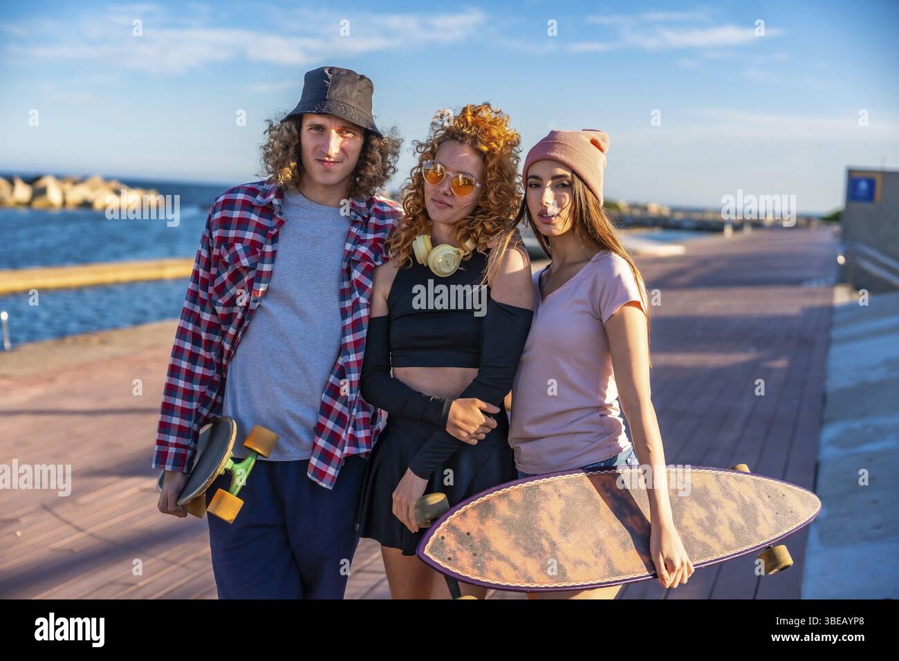Drei Freunde mit Skateboards entspannen sich am Meer und genießen das sonnige Wetter und die ungezwungene Atmosphäre im Freien auf einer hölzernen Promenade Stockfoto