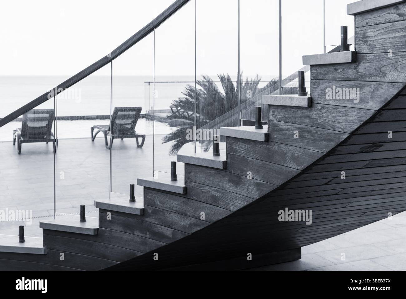 Elegante Holztreppe mit Glasgeländer und Blick auf eine Terrasse am Strand mit Liegestühlen und Meerblick, die moderne Architektur mit Küstenarchitektur verbindet Stockfoto