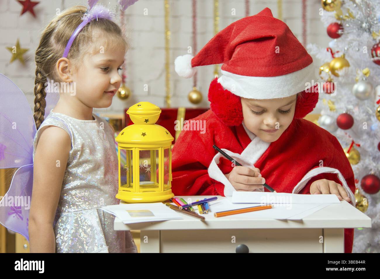 Ein Mädchen in einem roten Anzug Santa Claus Bleistifte am Tisch sitzen zieht gekleidet, nächsten ist ein Mädchen gekleidet wie ein Märchen mit dem Leuchter. Stockfoto