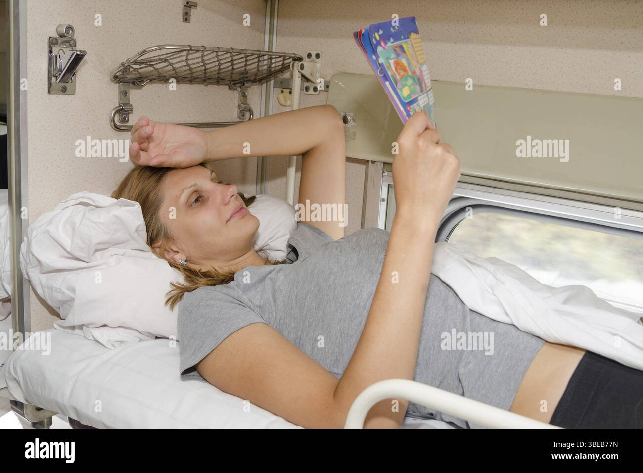 Das Mädchen im Zug auf dem Rücken auf dem obersten Regal und auf der Seite liegend, mit einem Lächeln Lesen einer Zeitschrift Stockfoto