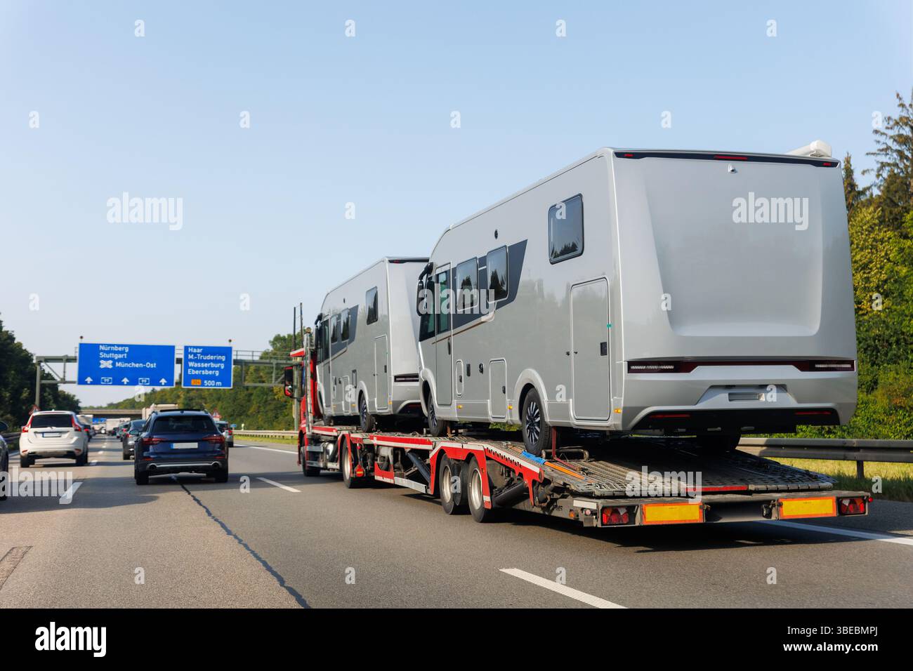Zwei neue moderne Wohnmobile transportierten Anhänger-LKW, befahrene deutsche Autobahn Stadtschilder. Die Fahrzeuge bewegen sich am sonnigen, blauen Himmel. Transporturlaub Stockfoto
