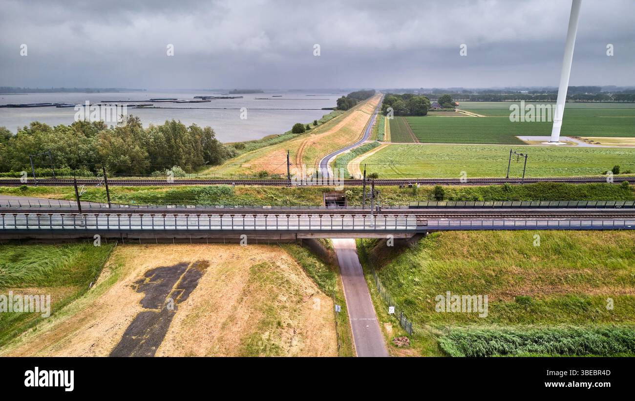 27. Mai 2025: Moerdijk-Niederlande: Aus der Vogelperspektive der Moerdijk-Eisenbahnbrücken über den Deich bei Hollands Diep. Grüne Flussufer, Fahrbahn, Viadukt Stockfoto