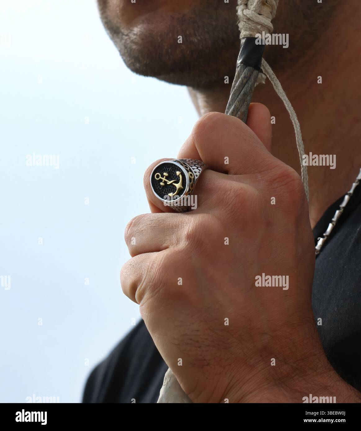 Nahaufnahme einer Matrosenhand, die einen silbernen Ring mit goldenem Anker trägt und ein Seil hält, das den Lebensstil der Seefahrt und maritimen Schmuck darstellt Stockfoto