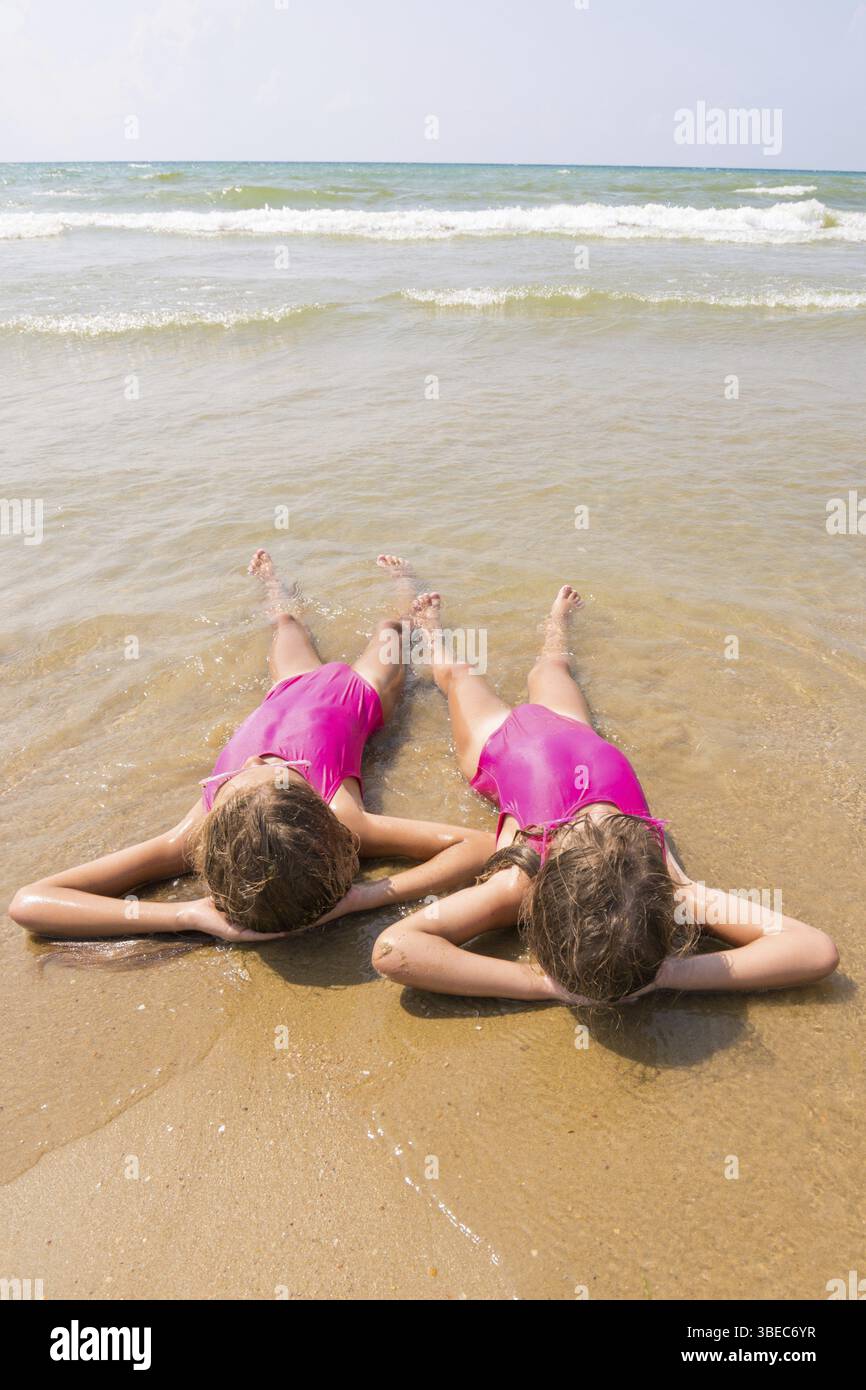 Zwei Mädchen liegen auf dem Rücken auf dem sandigen Strand auf halbem Weg in die wate Stockfoto