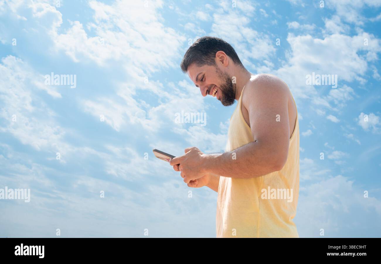Lachende junge Erwachsene Spanisch am Strand, SMS mit Handy und schönem sonnigen blauen Himmel Stockfoto