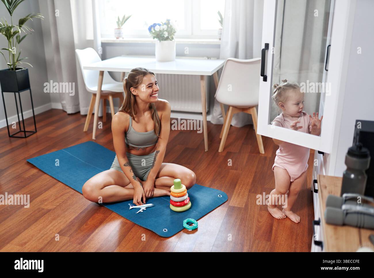 Eine fröhliche und fröhliche Mutter übt ihre Yoga-Routine zu Hause, während ihr liebenswertes Baby in der Nähe spielt und eine wunderbare Mischung aus Passform bietet Stockfoto