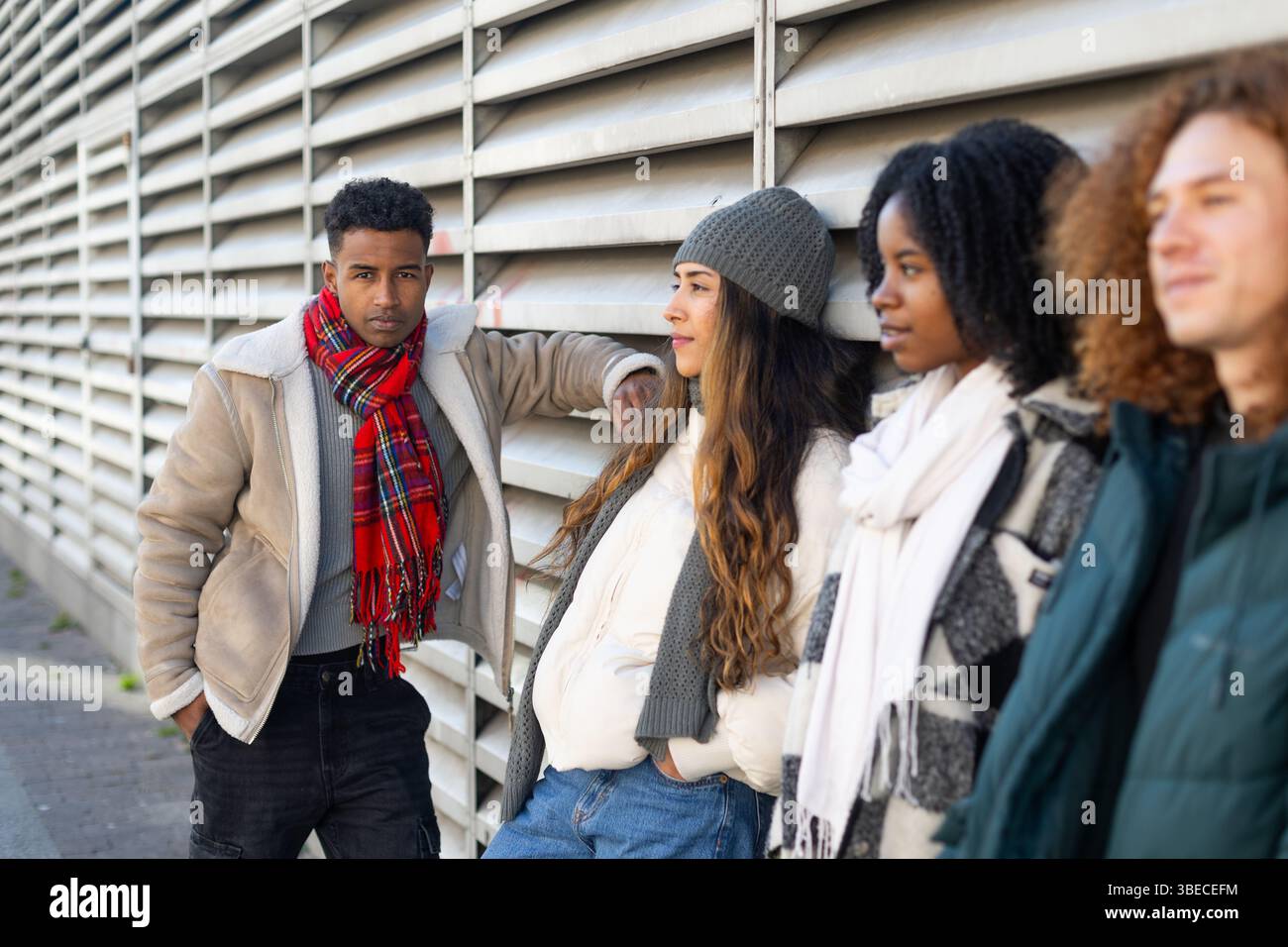 Vier verschiedene College-Studenten in Winterkleidung, die sich an eine Wand lehnen und zwischen den Unterrichtseinheiten eine Pause einlegen Stockfoto