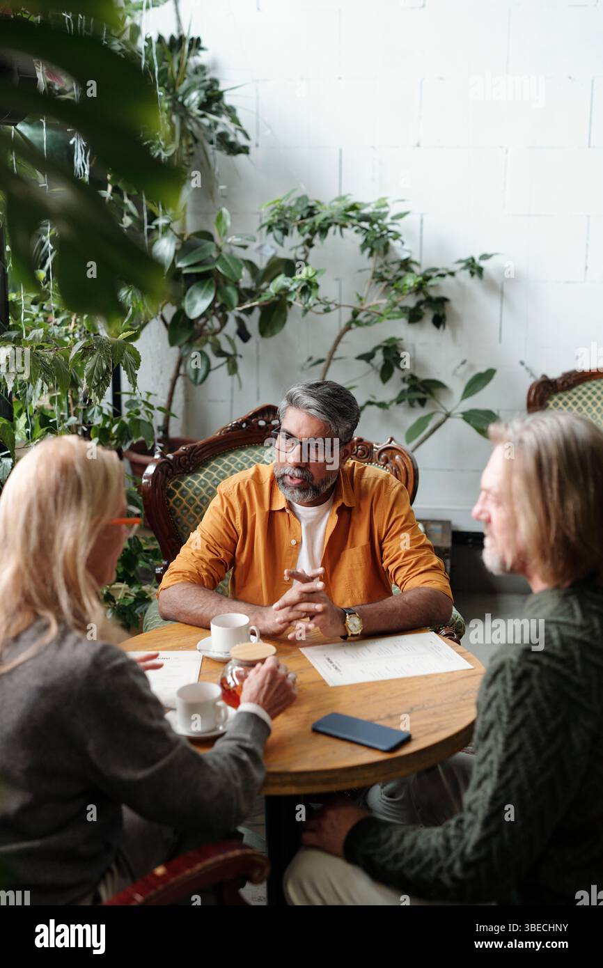 Eine Gruppe von drei Personen, die an einem runden Tisch sitzen und Pläne in gemütlicher Gartenatmosphäre diskutieren, umgeben von üppigen grünen Pflanzen, sieht entspannt aus und führt Gespräche Stockfoto