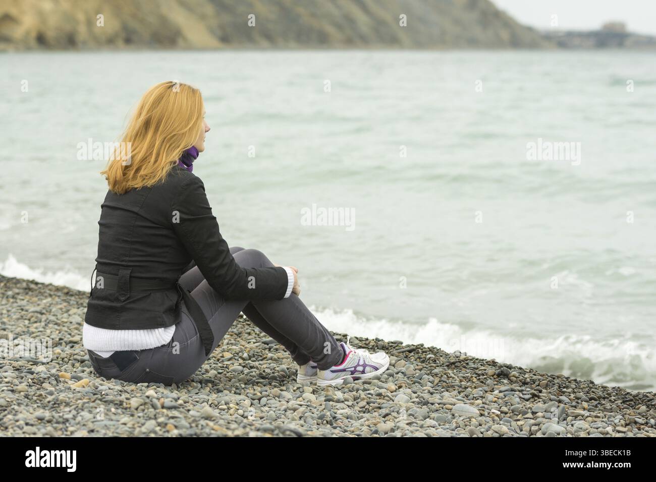 Mädchen in einem kühlen Tag am Strand sitzen und nachdenklich in die Ferne schauen Stockfoto