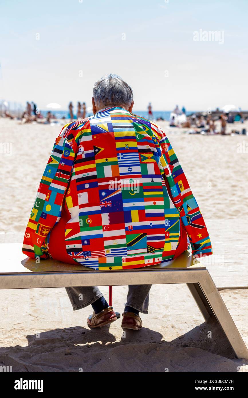 Spanien, Sitges, Ein Mann mit einer Jacke aus Fahnen der Welt, der am Strand sitzt Stockfoto