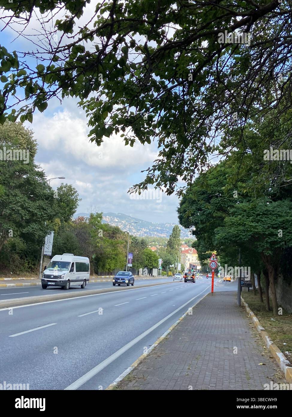 Von Bäumen gesäumte Stadtstraße mit Gehwegen und Fahrzeugen unter einem bewölkten blauen Himmel in Istanbul. - Smartphone-aufgenommenes Stockfoto