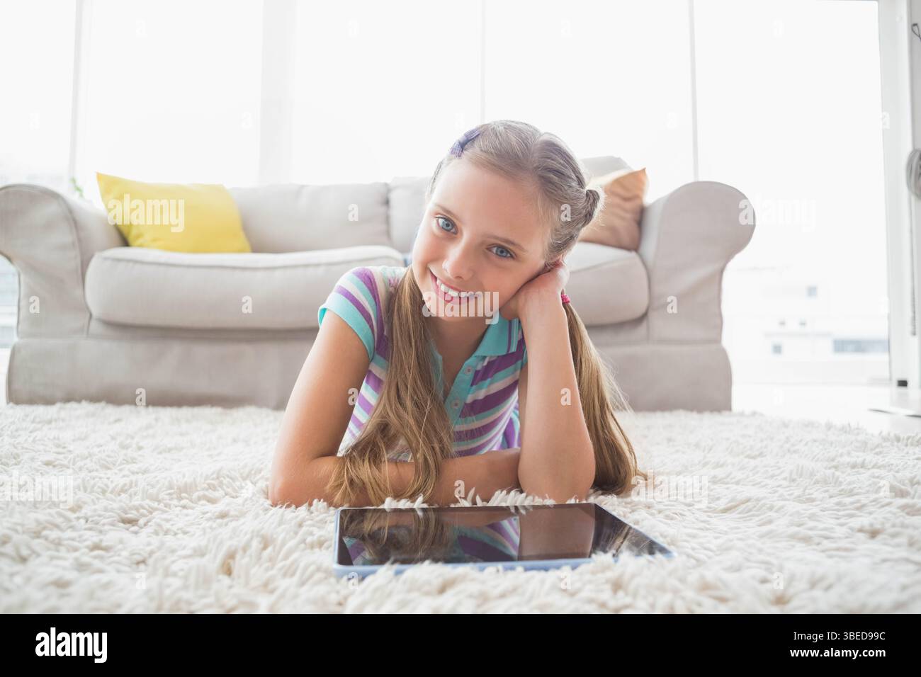 Schulmädchen, das auf einem weißen Teppich im hellen Wohnzimmer liegt und auf die Tablette blickt Stockfoto