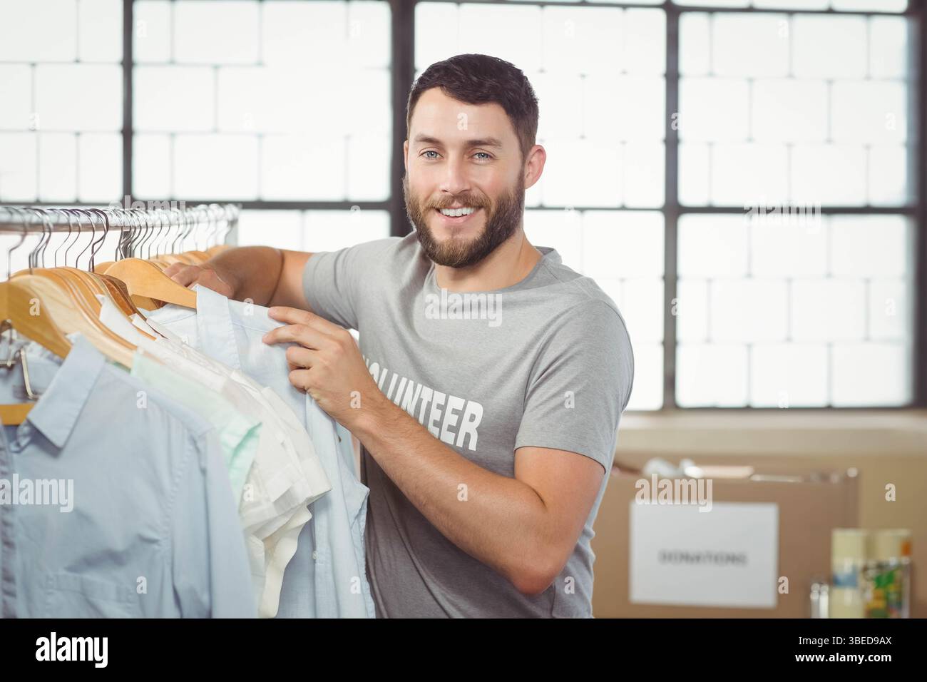 Männliche Freiwillige sortieren gespendete Hemden auf hölzernen Kleiderbügeln neben Spendenboxen im Lager Stockfoto