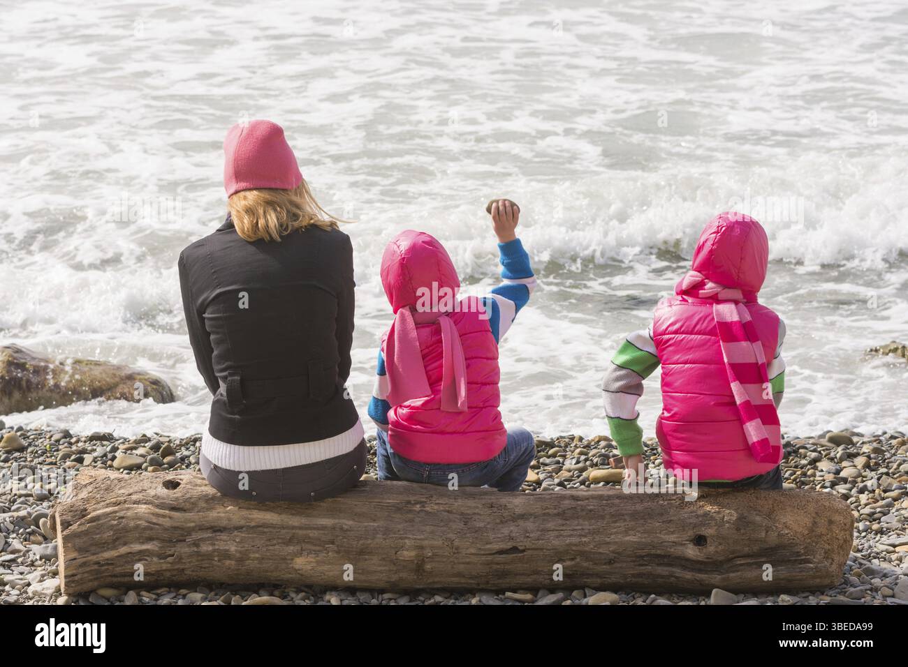 Zwei Mädchen und Frau sitzen auf einem Baumstamm am Strand und Steine ins Wasser Stockfoto