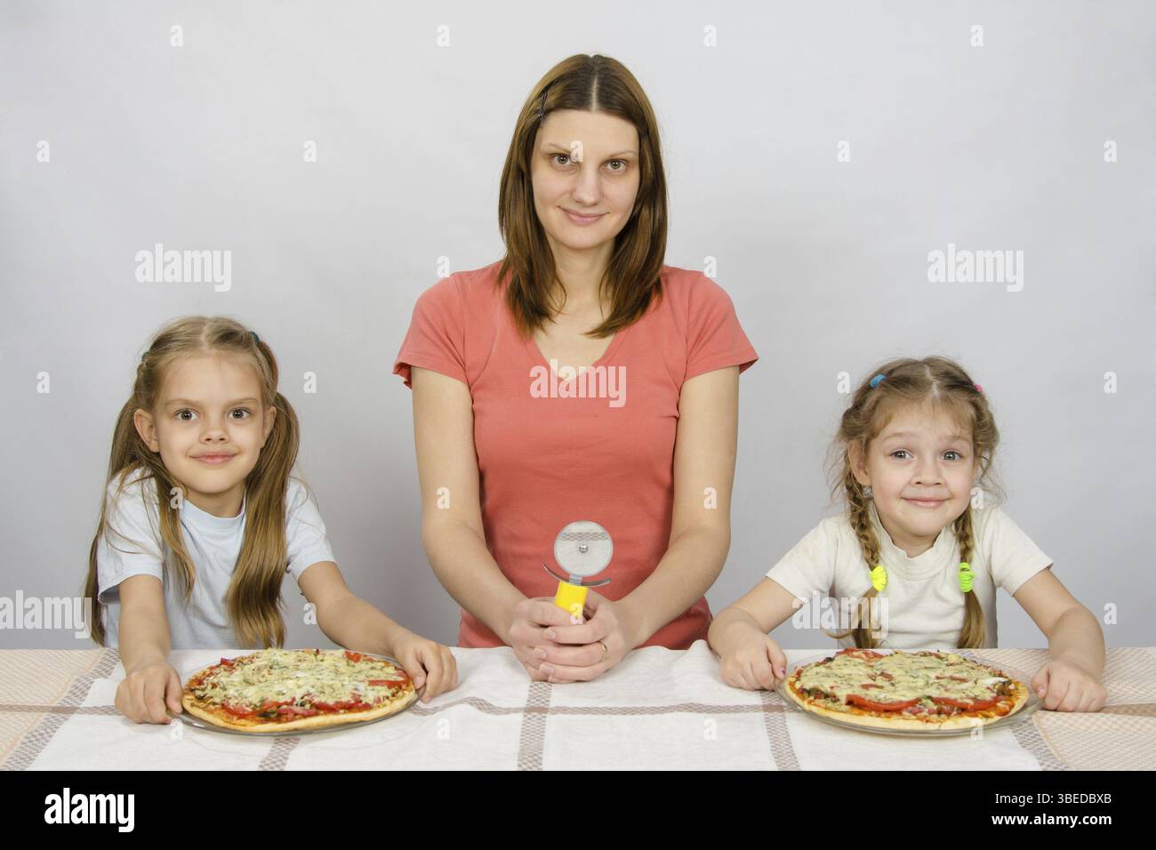 Am Tisch saß meine Mutter mit einem Messer für Pizza und zwei Töchter, denen zwei Pizzen Stockfoto