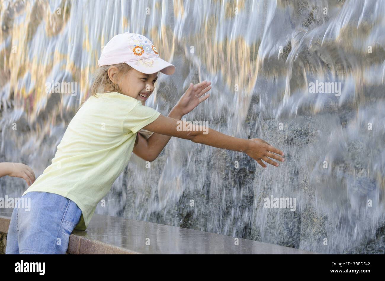 Fröhlich sechs Jahre altes Mädchen im Sommer Kleidung Hand versucht den Wasser künstliche Wasserfall zu erhalten Stockfoto