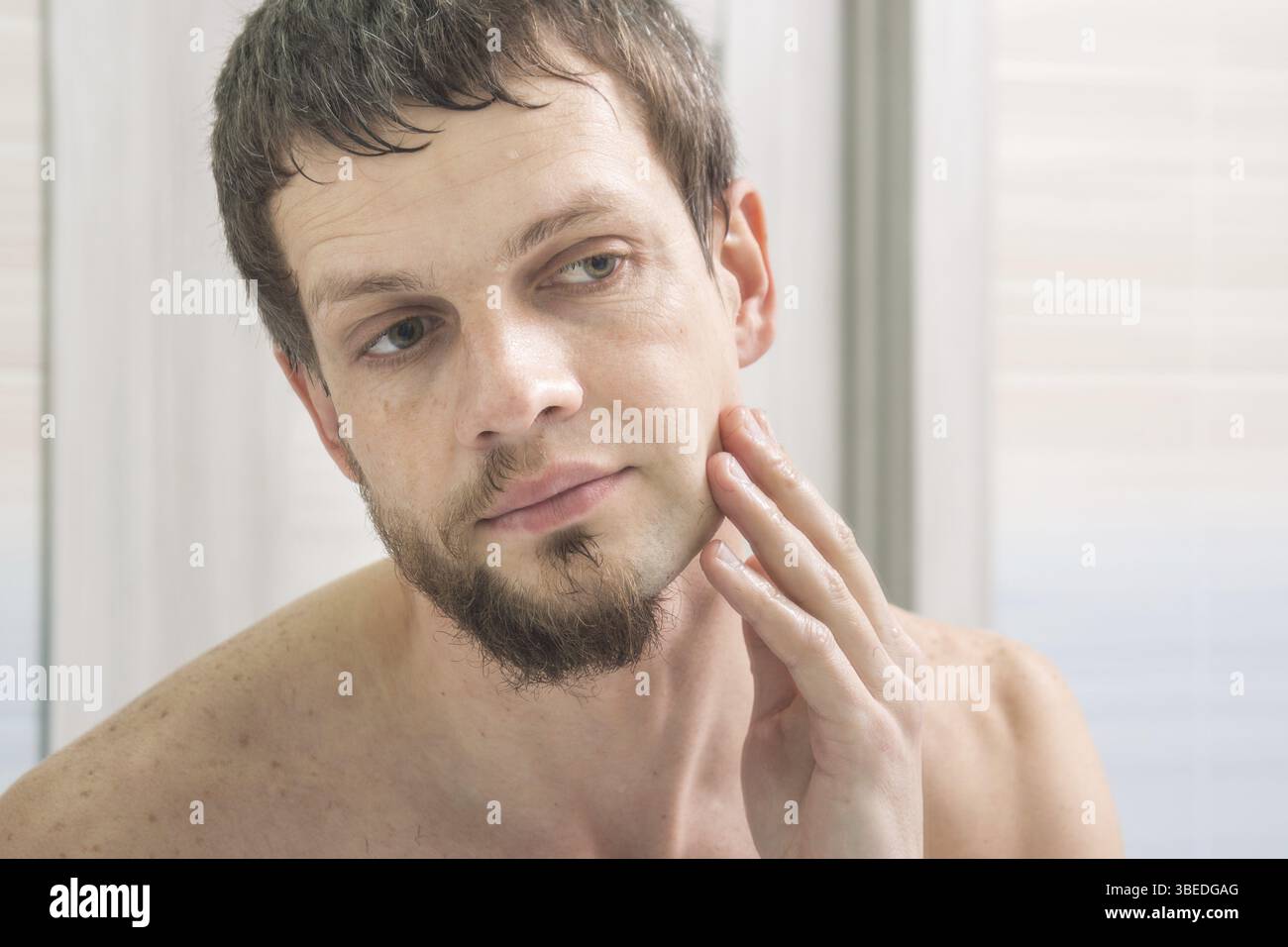 Der Kerl rasiert eine Hälfte von seinem Gesicht und sieht im Spiegel der Bewertung das Ergebnis Stockfoto