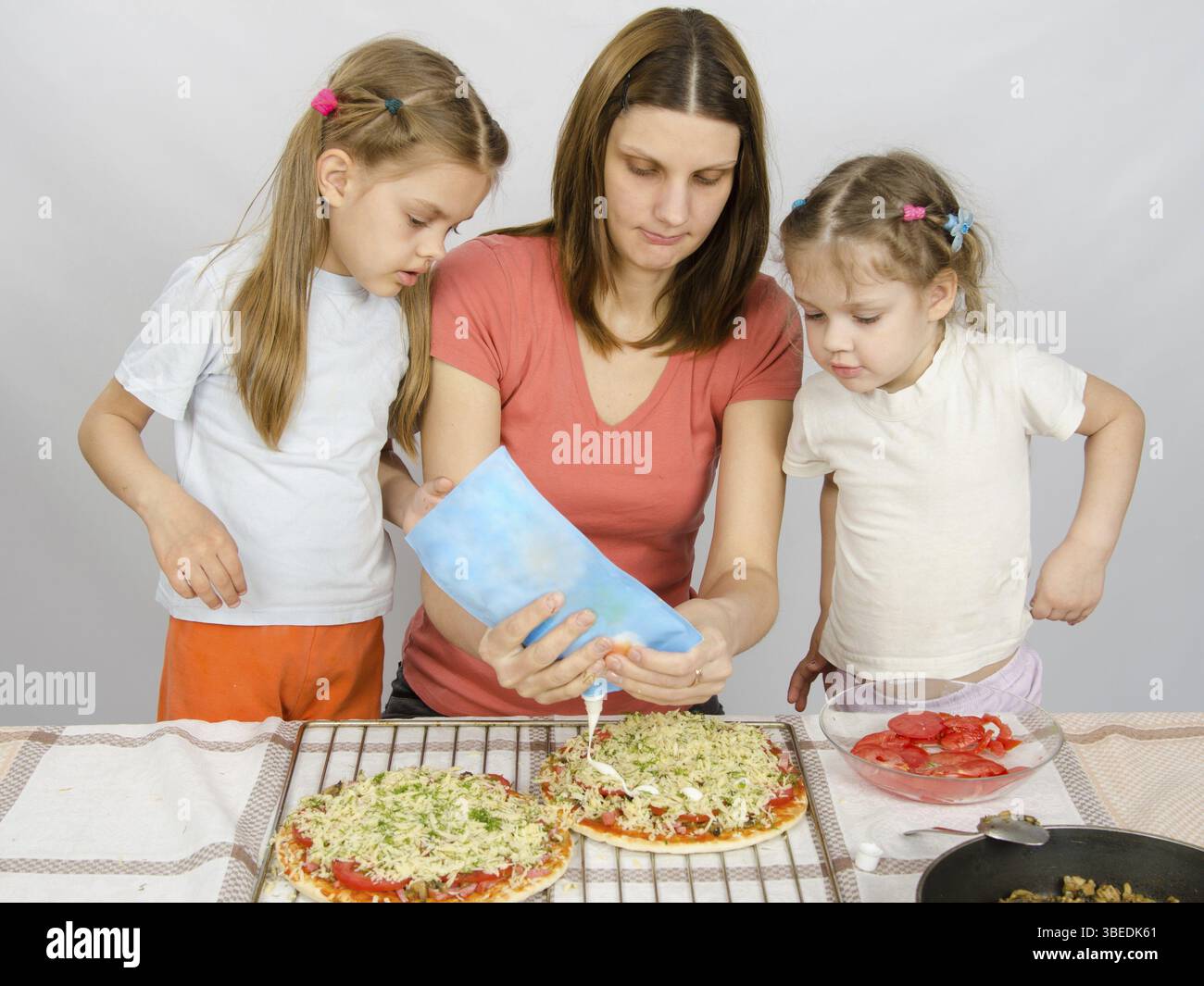 Zwei kleine Mädchen sind mit Interesse beobachten, wie die Mutter ist Mayonnaise Pizza gießen Stockfoto
