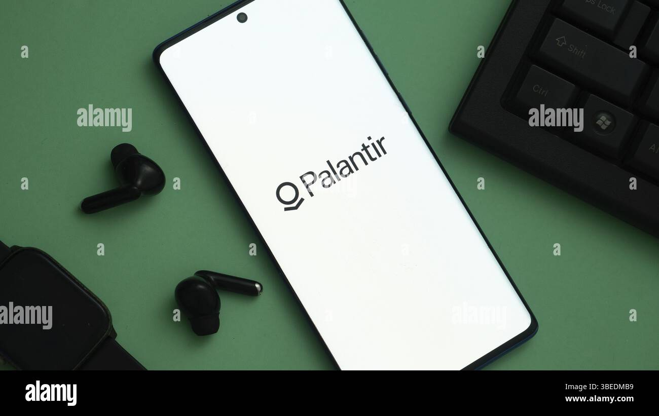 Palantir-Logo auf einem Smartphone-Bildschirm, umgeben von technischem Zubehör auf grüner Oberfläche Stockfoto