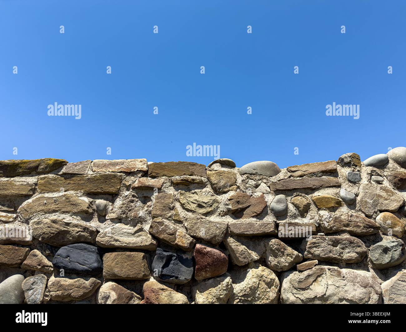 Alte rustikale Steinmauer mit unregelmäßigen Felsen und rauer Textur unter klarem blauem Himmel, Tageslicht, natürlichem Hintergrund für Design oder Architektur. Keine Personen. Stockfoto