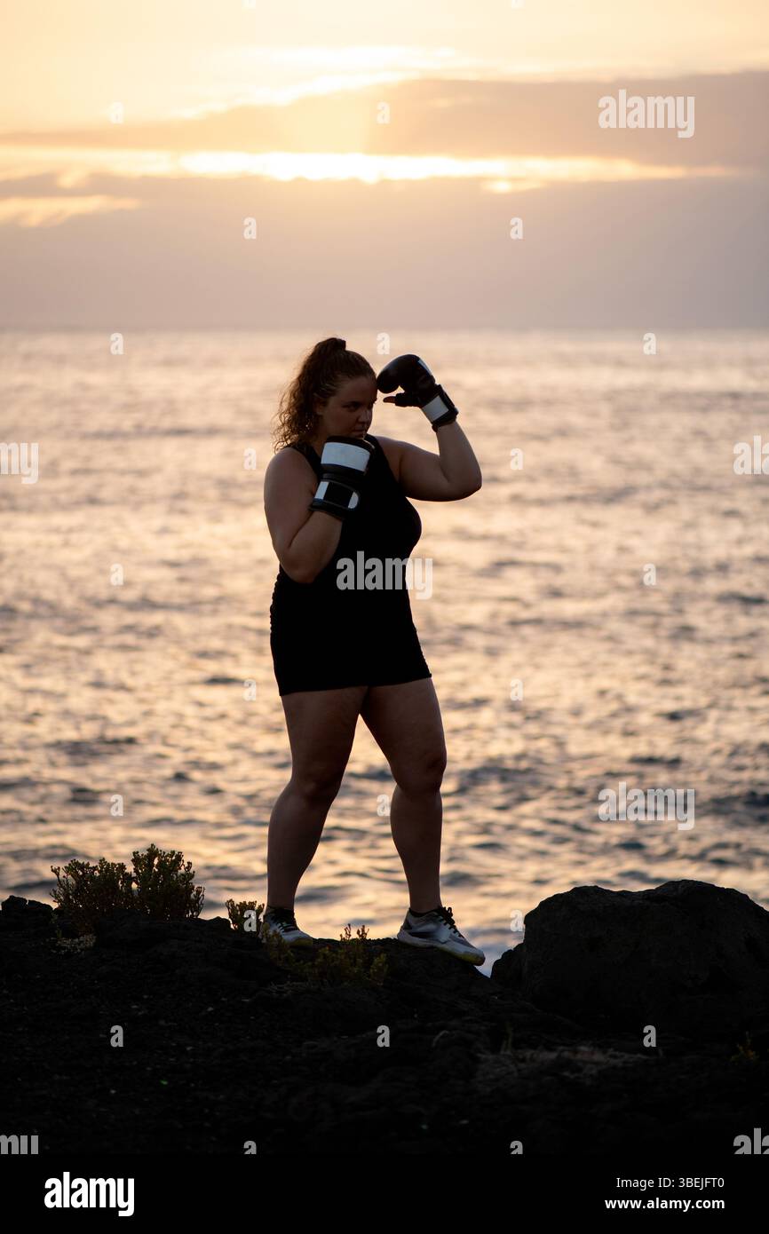 Entschlossene Frau, die ein ärmelloses schwarzes Workout-Outfit und Boxhandschuhe trägt und bei Sonnenuntergang auf schwarzen vulkanischen Felsen am Meer steht, Silhouetten Stockfoto