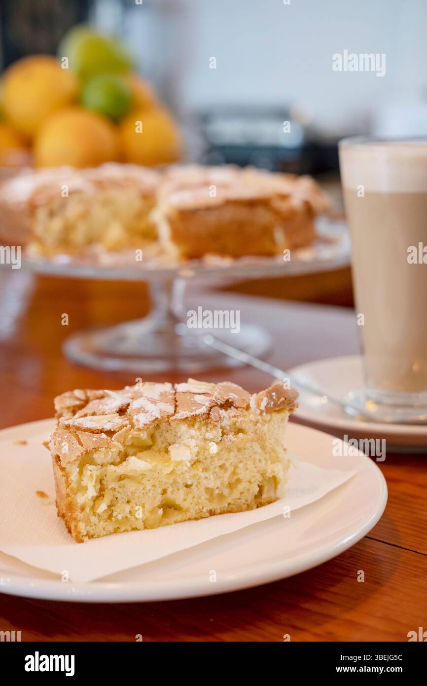 Ein frisch geschnittenes Stück hausgemachter Mandel- und Apfelkuchen auf einem Teller mit Serviette, ein großer Latte in einem hohen Glas und ein ganzer Kuchen auf einem Glasständer Stockfoto