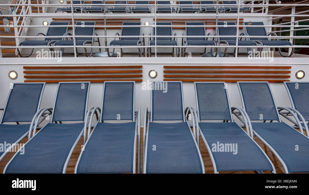 Ordentlich angeordnete Reihen leerer blauer Sonnenliegen auf einem Kreuzfahrtschiff-Deck, die symmetrische Ebenen und geometrische Geländerrahmen bilden Stockfoto