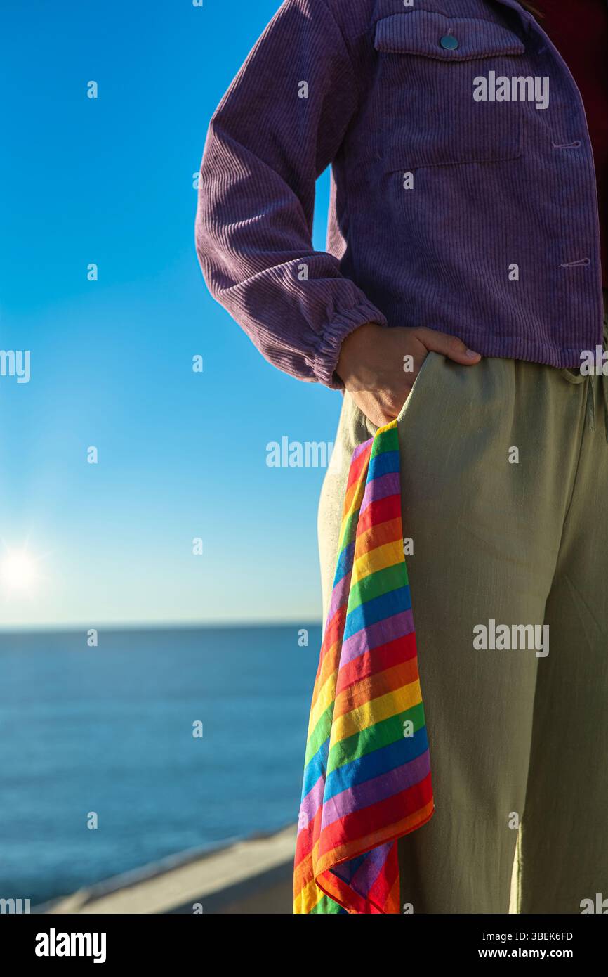 Vertikales Halbbild, das von der Schulter bis zum Knie aufgenommen wurde. Eine Hand liegt auf der Tasche der Hose, aus der ein Schal mit Regenbogenfarben, Symbol o Stockfoto