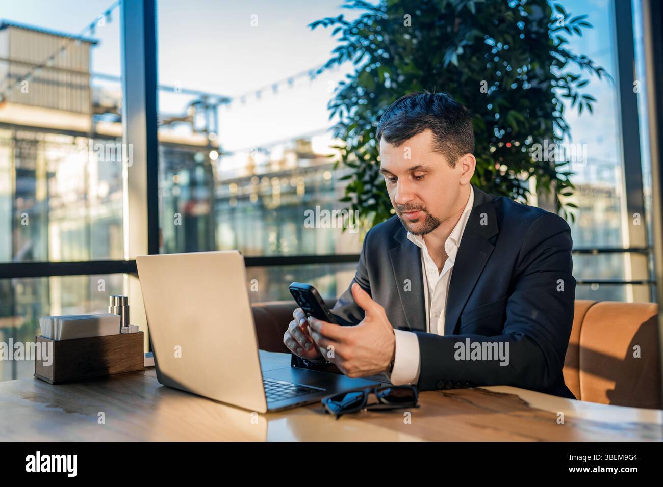 Ein Geschäftsmann im Anzug arbeitet mit einem Laptop an einem Kaffeetisch. Die Umgebung ist entspannt, und das Tageslicht strömt durch große Fenster und schafft so etwas Stockfoto