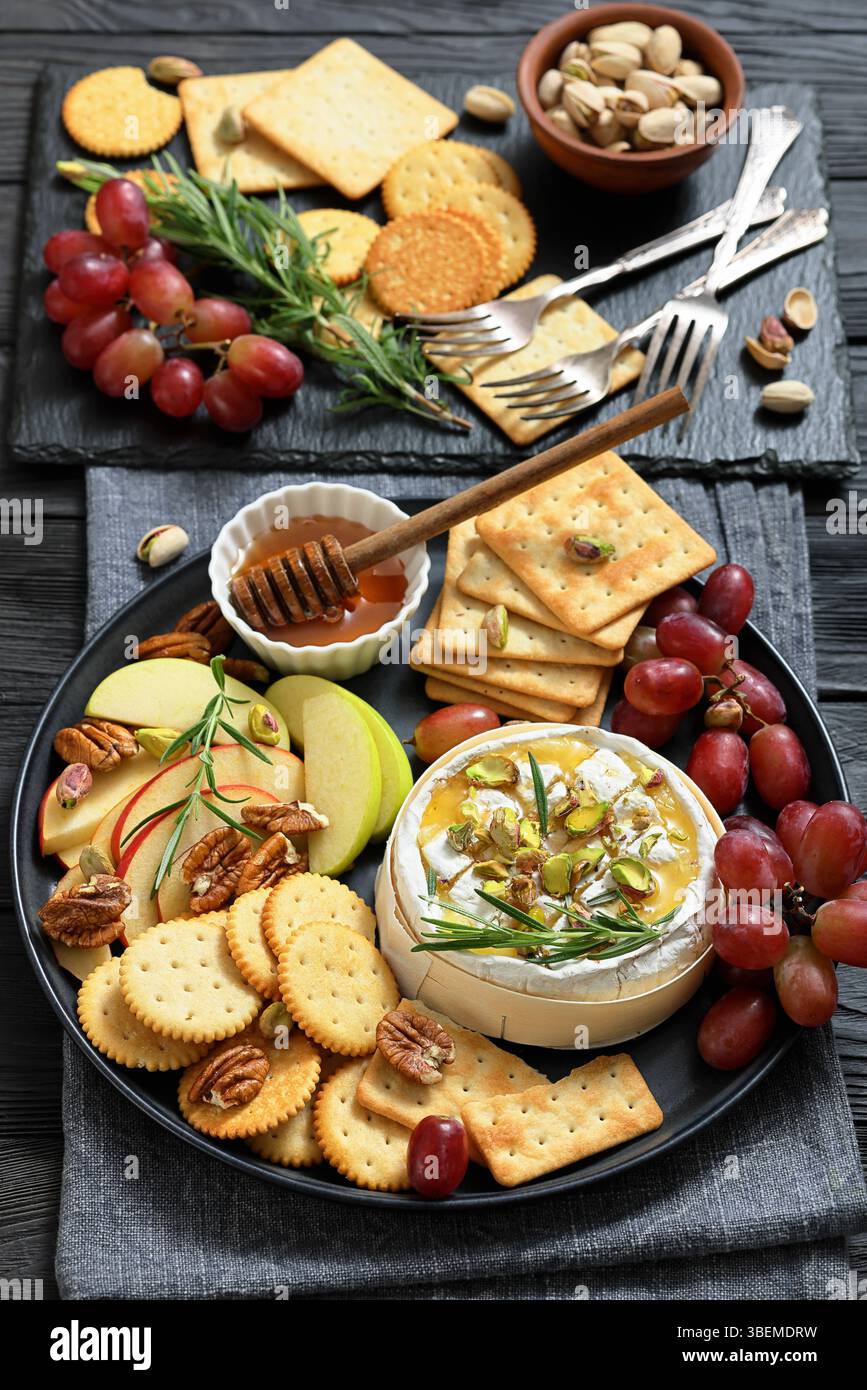 Camembert mit Honig und Pistazien, serviert mit Apfelscheiben, Trauben und Crackern auf schwarzem Holztisch mit Zutaten, echtes Lebensmittelfoto, kein KI-Gen Stockfoto