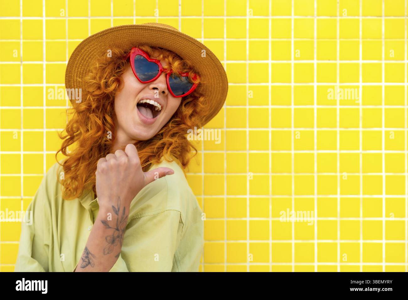 Fröhliche Influencerin zeigt mit ihrem Daumen, während sie lächelt, trägt einen Strohhut und eine herzförmige Sonnenbrille an einer gelb gekachelten Wand Stockfoto