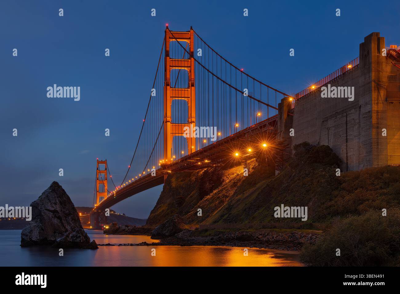 Die majestätische Golden Gate Hängebrücke erstreckt sich über ein ruhiges Gewässer, beleuchtet von warmen, leuchtenden Lichtern vor einem tiefblauen Abendhimmel. Stockfoto