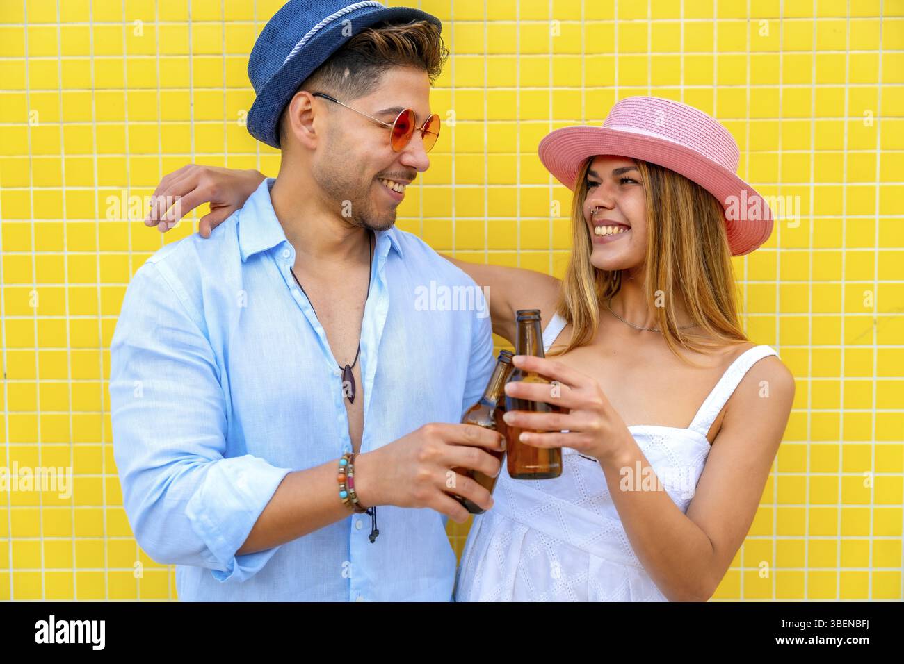 Junge, stilvolle Touristen, die mit Bierflaschen anstoßen, genießen gemeinsam den Sommerurlaub vor einer lebhaften, gelb gekachelten Wand Stockfoto