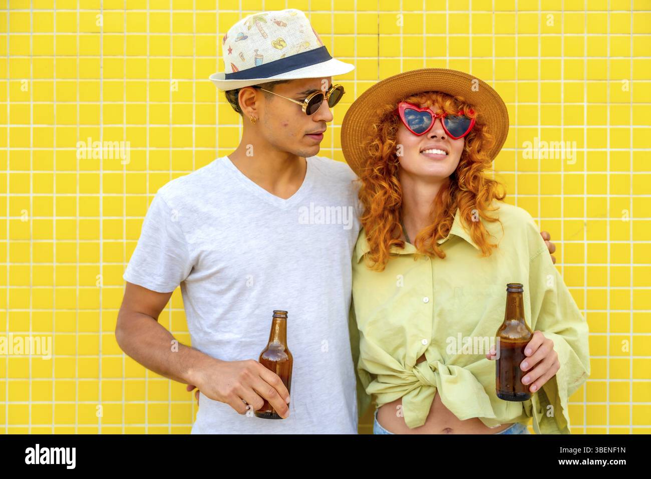 Ein junges Paar trägt Hüte und Sonnenbrille, hält Bierflaschen und lächelt fröhlich vor einer leuchtend gelben Wand, strahlt Glück aus Stockfoto
